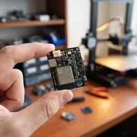 Papan dipegang di antara dua jari, dengan printer 3D di belakangnya